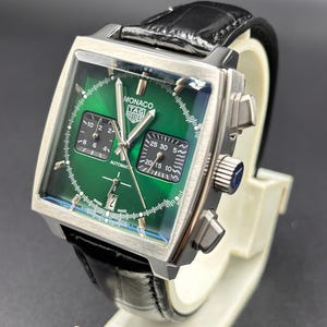 K&ouml;nnte beinhalten: Eine quadratische TAG Heuer Monaco Armbanduhr mit gr&uuml;nem Sonnenschliff-Zifferblatt, zwei schwarzen Hilfszifferbl&auml;ttern und einer Datumsanzeige auf der Sechs-Uhr-Position. Die Uhr hat silberfarbene Zeiger, Indizes, ein Edelstahlgeh&auml;use und ein schwarzes Lederarmband.