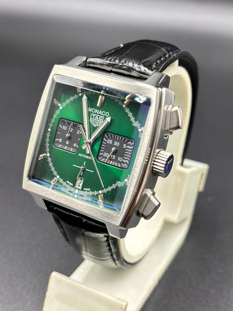 K&ouml;nnte beinhalten: Eine quadratische TAG Heuer Monaco Armbanduhr mit gr&uuml;nem Sonnenschliff-Zifferblatt, zwei schwarzen Hilfszifferbl&auml;ttern und einer Datumsanzeige auf der Sechs-Uhr-Position. Die Uhr hat silberfarbene Zeiger, Indizes, ein Edelstahlgeh&auml;use und ein schwarzes Lederarmband.