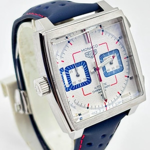 Może przedstawiać: Zegarek chronograf z kwadratową kopertą, srebrną teksturowaną tarczą, niebieskimi subtarczami i czerwonymi akcentami. Zegarek posiada napis MONACO HEUER oraz AUTOMATIC CHRONOGRAPH. Pasek wykonany jest z perforowanej granatowej sk&oacute;ry.