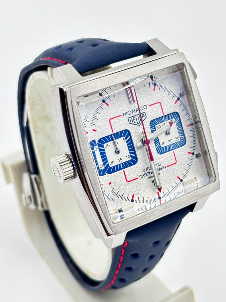 Może przedstawiać: Zegarek chronograf z kwadratową kopertą, srebrną teksturowaną tarczą, niebieskimi subtarczami i czerwonymi akcentami. Zegarek posiada napis MONACO HEUER oraz AUTOMATIC CHRONOGRAPH. Pasek wykonany jest z perforowanej granatowej sk&oacute;ry.