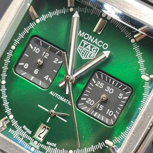 K&ouml;nnte beinhalten: Eine quadratische TAG Heuer Monaco Armbanduhr mit einem sonnengeschliffenen gr&uuml;nen Zifferblatt, silberfarbenen Zeigern und zwei schwarzen quadratischen Hilfszifferbl&auml;ttern. Die Uhr hat ein Datumsfenster auf sechs Uhr, wei&szlig;e Markierungen und den Schriftzug MONACO TAG HEUER.