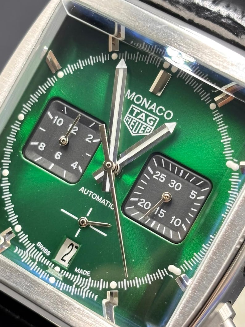 K&ouml;nnte beinhalten: Eine quadratische TAG Heuer Monaco Armbanduhr mit einem sonnengeschliffenen gr&uuml;nen Zifferblatt, silberfarbenen Zeigern und zwei schwarzen quadratischen Hilfszifferbl&auml;ttern. Die Uhr hat ein Datumsfenster auf sechs Uhr, wei&szlig;e Markierungen und den Schriftzug MONACO TAG HEUER.