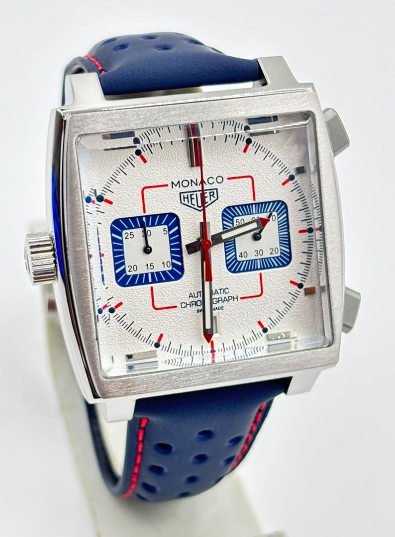 Może przedstawiać: Zegarek chronograf z kwadratową kopertą, srebrną tarczą, dwiema niebieskimi subtarczami i czerwonymi akcentami. Na tarczy widnieje napis MONACO HEUER oraz AUTOMATIC CHRONOGRAPH SWISS MADE. Posiada niebieski, perforowany sk&oacute;rzany pasek z czerwonymi przeszyciami.