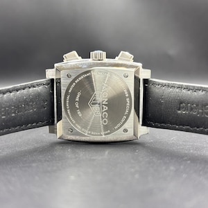 K&ouml;nnte beinhalten: Ein quadratisches Edelstahl-Uhrengeh&auml;use mit schwarzem Lederarmband. Der Geh&auml;useboden tr&auml;gt die Aufschrift Monaco, Heuer, 1969-1999 Special Edition, One of 169, Water Resistant 100 Meters und Swiss Made Since 1860 Sapphire Crystal.