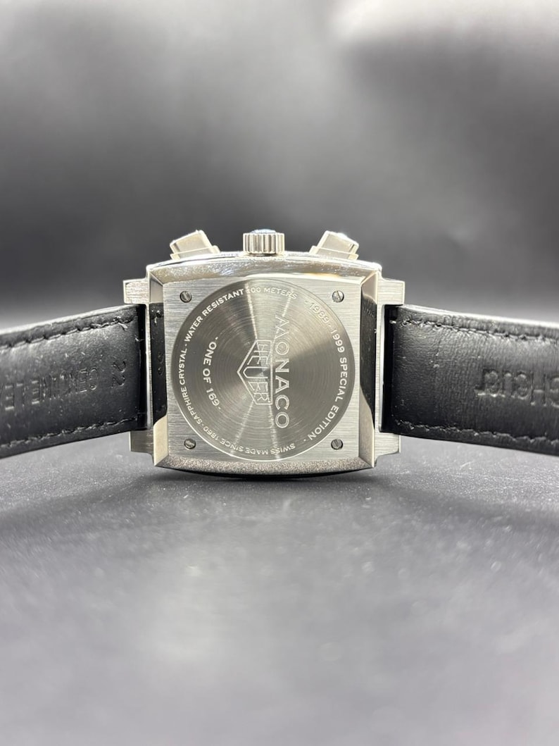 K&ouml;nnte beinhalten: Ein quadratisches Edelstahl-Uhrengeh&auml;use mit schwarzem Lederarmband. Der Geh&auml;useboden tr&auml;gt die Aufschrift Monaco, Heuer, 1969-1999 Special Edition, One of 169, Water Resistant 100 Meters und Swiss Made Since 1860 Sapphire Crystal.