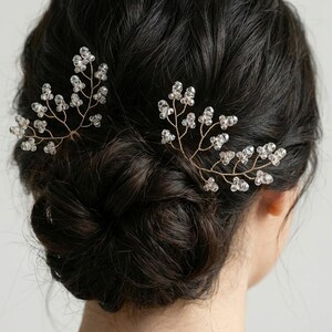 Könnte beinhalten: Zwei zarte Haarnadeln, jede mit einem goldfarbenen Drahtdesign mit klaren, funkelnden Kristallakzenten. Die Nadeln sind in einem floralen Muster angeordnet und verleihen dem dunklen Haar einen Hauch von Eleganz.