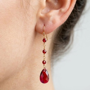 Puede incluir: Pendientes colgantes dorados con una serie de pequeñas cuentas rojas redondas que conducen a una gran gema roja facetada en forma de lágrima. Los pendientes se muestran en una oreja, destacando el diseño y el color.