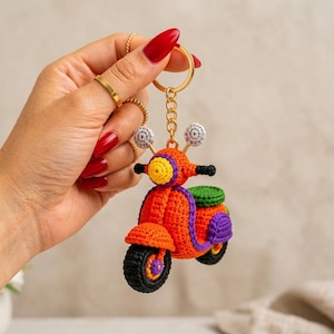 Può includere: Un portachiavi a forma di scooter realizzato all'uncinetto con corpo arancione vivace, sella verde, dettagli viola e ruote nere. Lo scooter in miniatura è fissato a un anello e una catenella in metallo dorato, con dettagli in filato lavorato.