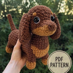 Puede incluir: Un juguete de perro salchicha tejido a ganchillo con hilo marrón oscuro y tostado claro. El peluche tiene grandes ojos de seguridad de plástico negro, orejas largas y caídas, y una cola corta. Un sello circular muestra el texto PDF PATTERN.