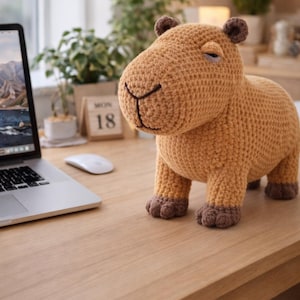 Puede incluir: Un peluche de carpincho tejido a crochet en tonos beige y marrón. El animal hecho a mano tiene una cara sonriente, orejas pequeñas y cuatro patas cortas. Está sobre un escritorio de madera cerca de un portátil y un calendario.
