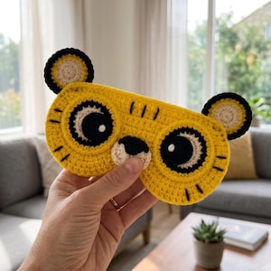 May include: A yellow crochet sleep mask shaped like a tiger face with black and white circular eyes, a small black nose, and two rounded ears with black borders. The mask features embroidered black whiskers and stripes on the yellow yarn surface.