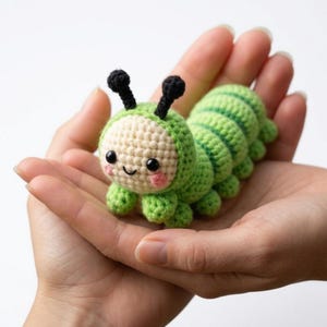 May include: A crocheted caterpillar toy in shades of green and cream, with black antennae and rosy cheeks. The handmade plushie has a smiling face and is held in open hands. The caterpillar's body is segmented with a striped pattern.
