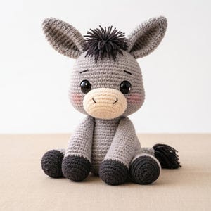 Puede incluir: Un peluche de burro hecho a mano con ganchillo en tonos grises, negros y crema. El burro tiene ojos negros grandes y expresivos, un hocico color crema y un pequeño mechón de hilo negro para la melena. El peluche está sentado con las piernas extendidas.