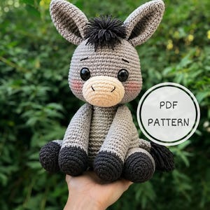 May include: A crocheted donkey toy with grey yarn, a beige muzzle, black hooves, and a black yarn tuft on its head. The donkey has large black safety eyes and pink blush details on its cheeks. A white circular graphic displays the text PDF PATTERN.