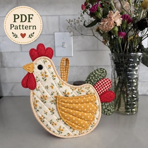 May include: A quilted fabric chicken-shaped potholder featuring a cream body with a yellow floral print, a red comb and wattle, a yellow polka-dot wing, and a tail with green leaf and red gingham patterns. A yellow gingham loop is attached. Text reads PDF Pattern.