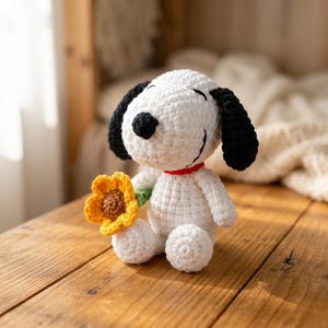 Op de afbeelding: Een wit gehaakt hondenfiguurtje met zwarte oren en een zwarte neus, met een dunne rode halsband om de nek. De hond zit rechtop en houdt een kleine, felgele gehaakte bloem met een bruin hartje vast in zijn voorpoot.