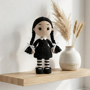 Könnte beinhalten: Eine handgefertigte Häkelpuppe mit langen, schwarzen Zöpfen, einem schwarzen Kleid mit weißem Kragen und schwarz-weiß gestreiften Strümpfen. Die Puppe hat schwarze Knopfaugen, weiße Schleifen an den Zöpfen und schwarze Schuhe mit kleinen Knöpfen.
