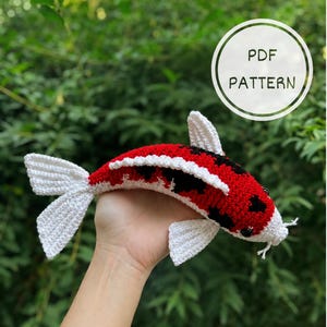 May include: A crocheted koi fish toy featuring a white, red, and black color pattern with white fins and tail. The fish has small black eyes and a white mouth area. A circular white graphic overlay in the upper right corner displays the text PDF PATTERN.