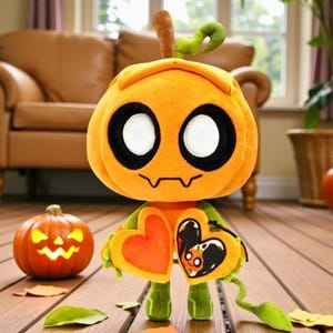 Peut inclure: Un personnage en peluche en forme de citrouille avec un visage souriant, de grands yeux blancs et des contours noirs. Il a des bras et des jambes verts et tient deux objets en forme de cœur. Une petite citrouille sculptée et des feuilles sont sur la surface en bois.
