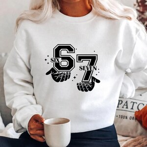 May include: White sweatshirt with a black graphic design featuring the numbers "6" and "7" with the words "SIX" and "SEVEN" above and below the numbers. The numbers are held by two hands.