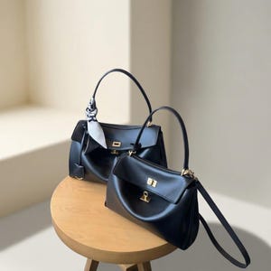 Könnte beinhalten: Zwei schwarze Lederhandtaschen mit goldfarbenen Verschlüssen und Henkeln stehen auf einem kleinen Holztisch. Eine Tasche hat ein dekoratives Tuch. Die Taschen haben ein klassisches, strukturiertes Design.