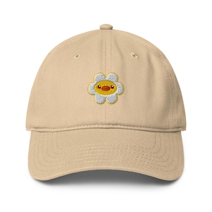 May include: A tan baseball cap with a curved brim. The cap features an embroidered design of a yellow and white flower with a small, round, red-orange face in the center. The cap is made of a soft fabric.