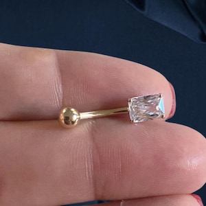 May include: A gold-colored belly button ring with a rectangular, clear gemstone and a small, round ball on the opposite end. The jewelry is held between fingers against a dark blue background.