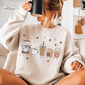 May include: A beige crewneck sweatshirt featuring a graphic print of various coffee-themed characters including an espresso machine, a mug, an iced coffee, a coffee bag, and a paper cup, all surrounded by small coffee beans and colorful stars.
