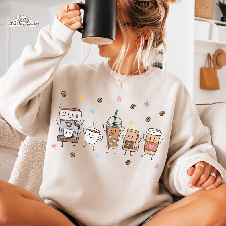 May include: A beige crewneck sweatshirt featuring a graphic print of various coffee-themed characters including an espresso machine, a mug, an iced coffee, a coffee bag, and a paper cup, all surrounded by small coffee beans and colorful stars.