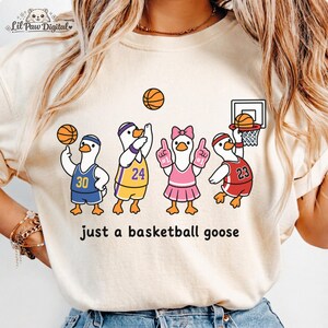Puede incluir: Camiseta color crema con una ilustración de dibujos animados de cuatro gansos jugando al baloncesto. Cada ganso lleva un uniforme de baloncesto con diferentes números. El texto "just a basketball goose" está impreso debajo.