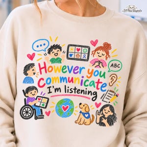 Puede incluir: Una sudadera beige con un gráfico colorido estilo crayón con el texto "However you communicate I'm listening." El diseño incluye ilustraciones de niños, una silla de ruedas, un perro, un globo terráqueo, corazones y símbolos de comunicación.