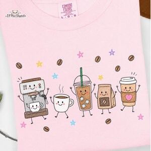 May include: A light pink t-shirt featuring a graphic print of five anthropomorphic coffee-themed characters including an espresso machine, a mug, an iced coffee, a coffee bag, and a paper cup, surrounded by coffee beans and colorful stars.