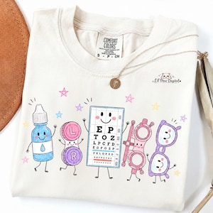 Puede incluir: Una camiseta de color crema con un estampado gráfico de artículos de cuidado ocular antropomórficos, que incluye un frasco de gotas azules, un estuche de lentes de contacto, una tabla optométrica con el texto EP TOZ, una gafa de prueba y gafas moradas.
