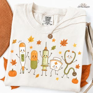 May include: A cream-colored t-shirt featuring a graphic print of five anthropomorphic medical items including a thermometer reading 98.6 F, a pill, a syringe, a nurse cap, and a stethoscope, surrounded by orange and yellow autumn leaves and stars.