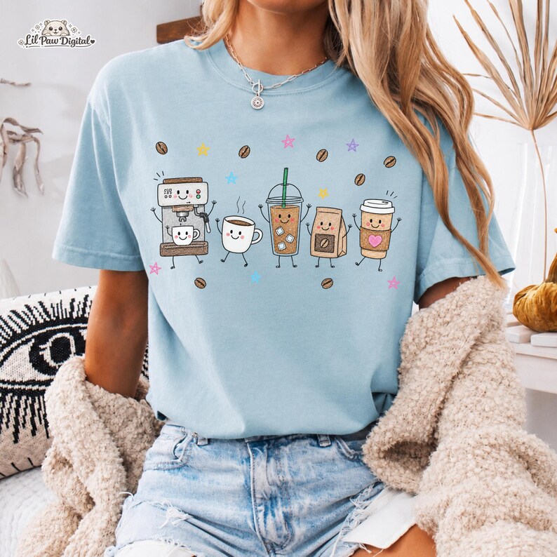 May include: A light blue t-shirt featuring a graphic print of five anthropomorphic coffee-themed items including an espresso machine, a mug, an iced coffee, a coffee bag, and a paper cup, surrounded by small coffee beans and colorful stars.