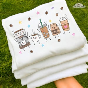 May include: A white sweatshirt featuring a colorful graphic print of anthropomorphic coffee items including an espresso machine, a mug, an iced coffee, a coffee bag, and a paper cup with a pink heart, surrounded by coffee beans and stars.