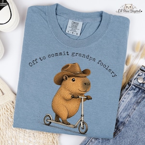 May include: A blue t-shirt featuring a graphic of a brown capybara wearing a brown cowboy hat and riding a silver kick scooter. The text above the capybara reads, Off to commit grandpa foolery. The shirt has a Comfort Colors brand neck label.