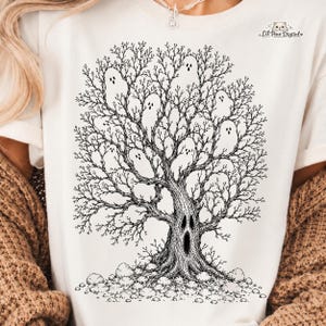 Op de afbeelding: Een crèmekleurig t-shirt met een zwarte inktillustratie van een boom zonder bladeren met een gezicht in de stam en meerdere spookfiguren in de takken. De tekst Lil Paw Digital staat in de rechterbovenhoek gedrukt.