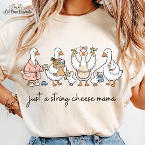 Puede incluir: Camiseta color crema con una ilustración caprichosa de gansos. Los gansos se representan en varios escenarios, como usando una bata, sosteniendo comestibles y llevando plátanos. El texto "just a string cheese mama" está escrito en cursiva debajo.