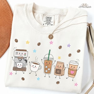 May include: A cream-colored t-shirt featuring a graphic print of five anthropomorphic coffee-themed characters, including an espresso machine, a mug, an iced coffee, a coffee bag, and a paper cup, surrounded by coffee beans and colorful stars.