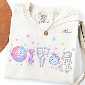 Op de afbeelding: Een crèmekleurig t-shirt met een kleurrijke afbeelding van antropomorfe fysiotherapie-apparatuur, waaronder een oefenbal, een halter, krukken, een ijszak en een pulsoximeter, omringd door kleine roze, gele en blauwe sterren.
