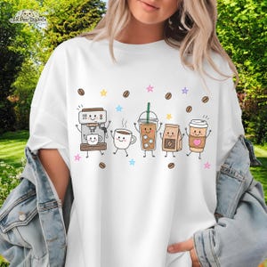 May include: A white t-shirt featuring a graphic print of five anthropomorphic coffee-themed characters including an espresso machine, a mug, an iced coffee, a coffee bag, and a paper cup, surrounded by small coffee beans and colorful stars.