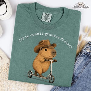 Könnte beinhalten: Ein salbeigrünes T-Shirt mit einer Grafik eines braunen Capybaras, das einen braunen Cowboyhut trägt und auf einem silbernen Tretroller fährt. Der Text "Off to commit grandpa foolery" ist in Weiß über der Capybara-Illustration aufgedruckt.