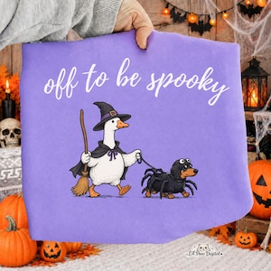 May include: A light purple t-shirt featuring a white graphic of a goose wearing a witch hat and cape while holding a broom, walking a dachshund dressed in a black spider costume. The text "off to be spooky" is printed in white above the graphic.