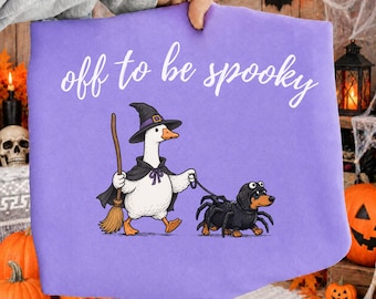 May include: A light purple t-shirt featuring a white graphic of a goose wearing a witch hat and cape while holding a broom, walking a dachshund dressed in a black spider costume. The text "off to be spooky" is printed in white above the graphic.