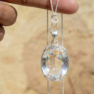 Puede incluir: Un collar con colgante de cristal transparente y facetado con una cadena de plata. El colgante grande, de forma ovalada, refleja un arcoíris de colores. El collar está sostenido por una mano sobre un fondo borroso de color marrón claro.