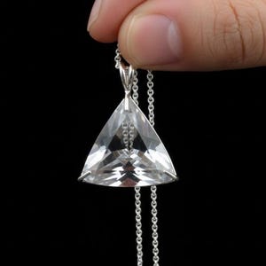 May include: A clear, triangular pendant necklace with a silver chain. The pendant is faceted, catching the light. The necklace is held against a black background, highlighting the jewelry's sparkle and design.
