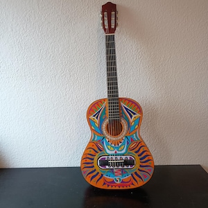 Può includere: Chitarra acustica con un design vibrante e colorato. La chitarra presenta un corpo arancione dipinto con motivi geometrici blu, gialli e viola. Il manico e la paletta sono marrone scuro. La chitarra è appoggiata su una superficie nera.