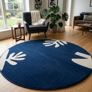 Könnte beinhalten: Ein runder, marineblauer Teppich mit einem cremefarbenen Blattmuster. Der Teppich liegt auf einem Holzboden in einem Wohnzimmer, mit einem grauen Sessel und einer Topfpflanze im Hintergrund. Der Teppich hat einen Durchmesser von ca. 200 cm.