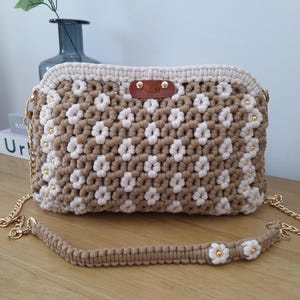 Puede incluir: Bolso bandolera de macramé hecho a mano en color beige y crema, con un diseño floral. El bolso tiene una correa de cadena dorada y un asa de macramé a juego con detalles de flores. Una pequeña etiqueta de cuero está adjunta.
